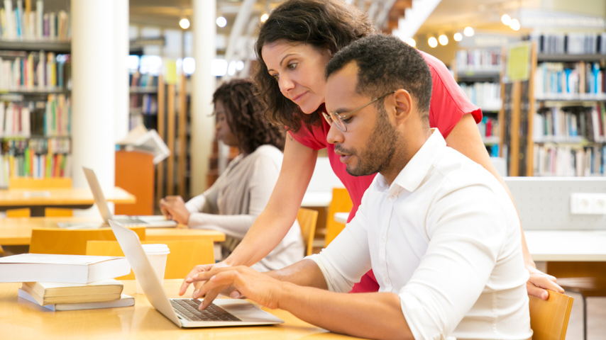 Interior de una biblioteca, estanterías llenas de libros, y en primer término, dos personas trabajando delante de un ordenador
